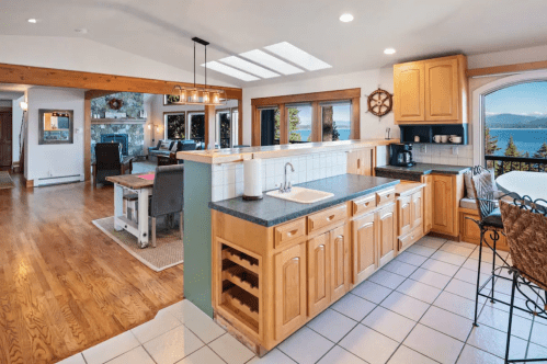 Bright kitchen with wooden cabinets, a sink, and a view of a lake, connected to a cozy living area with large windows.